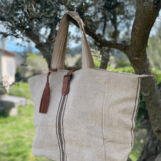 Sac cabas en toile beige avec anses et pompon en cuir, suspendu à un arbre en extérieur